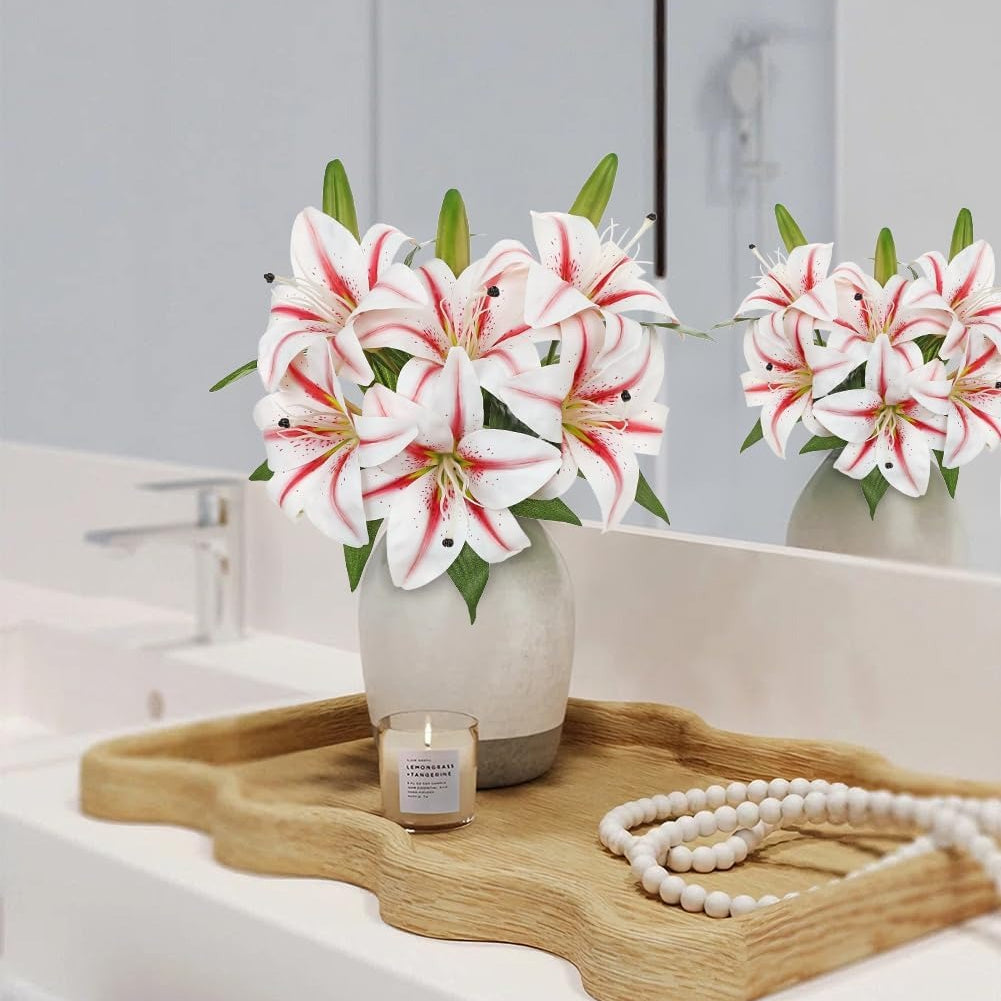 White lilies with red stripes in a light gray ceramic vase, styled on a wooden tray with a candle and a pearl necklace on a white bathroom counter.