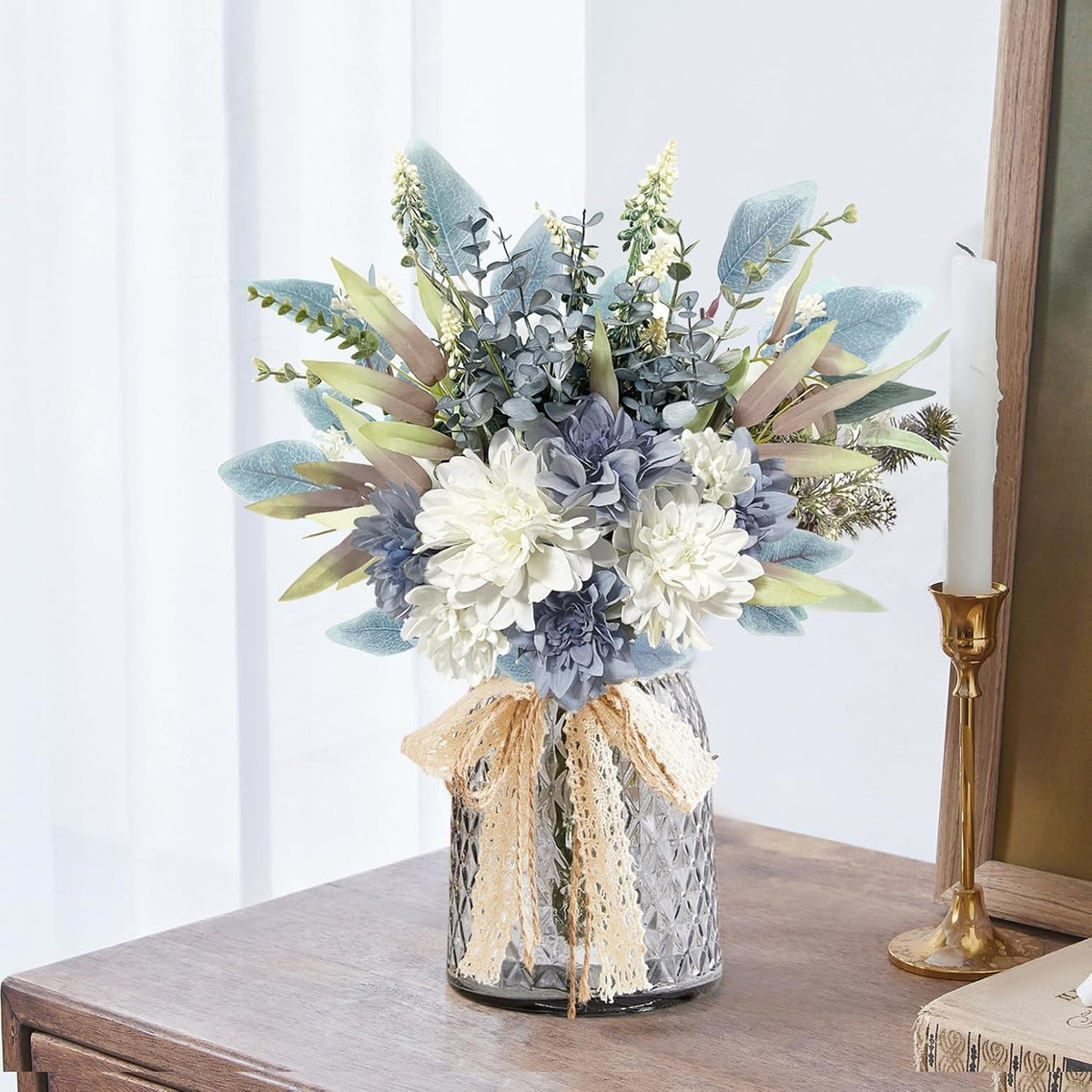 Decorative floral arrangement in a glass vase on a wooden surface with a white curtain background.