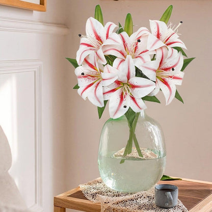 A bouquet of white lilies with red streaks and green leaves arranged in a clear glass vase, placed on a wooden table in a softly lit interior setting.