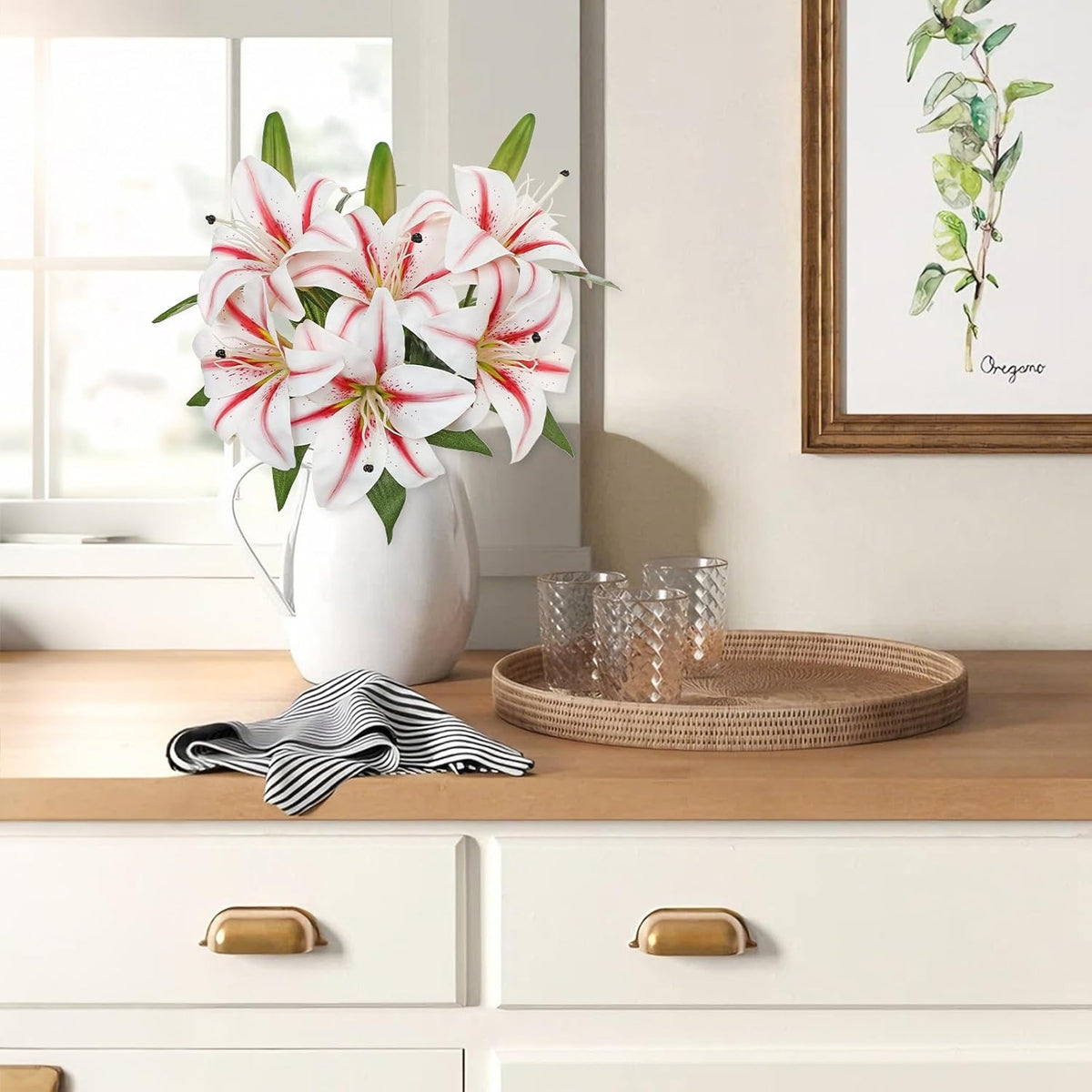 A bouquet of white lilies with red stripes in a white pitcher vase sits on a light wood countertop beside two clear textured drinking glasses and a woven round tray. The scene is styled in a bright kitchen or dining area with white cabinets, a striped cloth, and framed wall art.