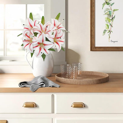 A bouquet of white lilies with red stripes in a white pitcher vase sits on a light wood countertop beside two clear textured drinking glasses and a woven round tray. The scene is styled in a bright kitchen or dining area with white cabinets, a striped cloth, and framed wall art.
