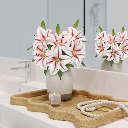 White lilies with red stripes in a light gray ceramic vase, styled on a wooden tray with a candle and a pearl necklace on a white bathroom counter.