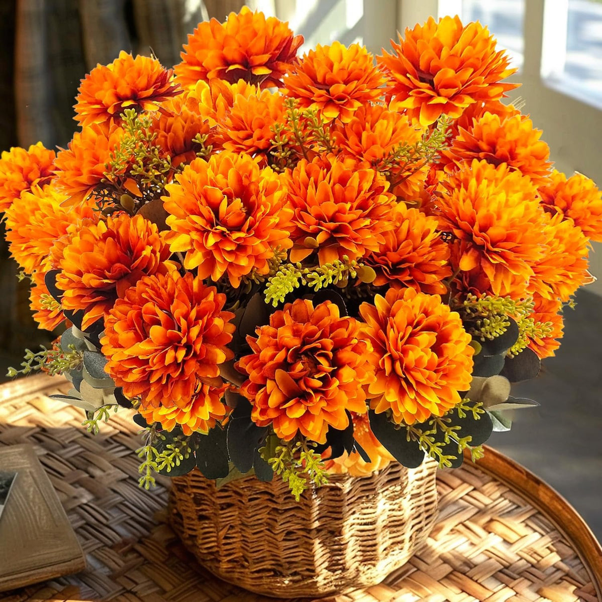 A large arrangement of bright orange artificial flowers with green and yellow filler stems, displayed in a woven basket on a round woven table.