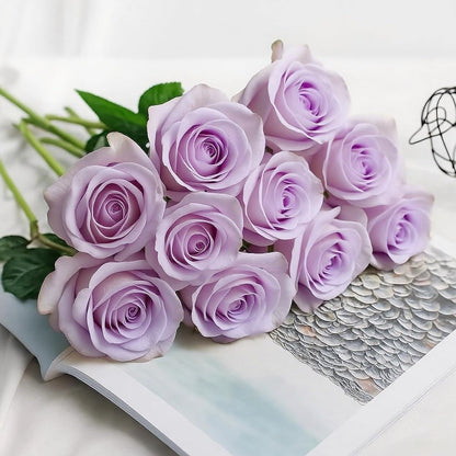 A bouquet of light purple/lavender artificial roses with green stems, arranged on an open book over a white fabric background.