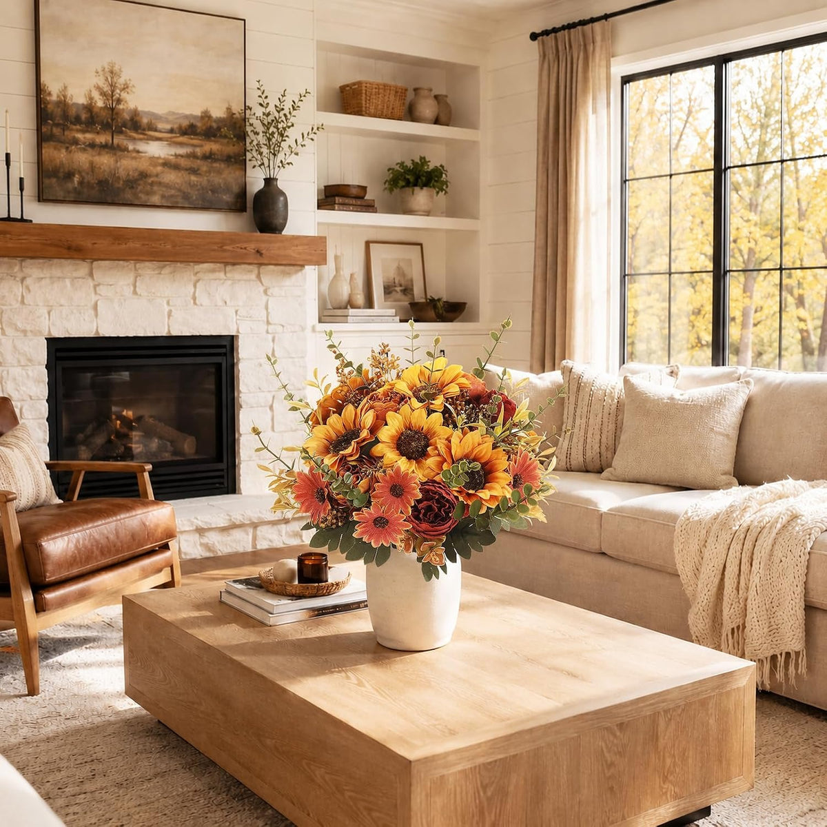 A styled living room featuring a large wooden coffee table in the foreground with a white vase of sunflowers, set in front of a beige sofa and a white brick fireplace.