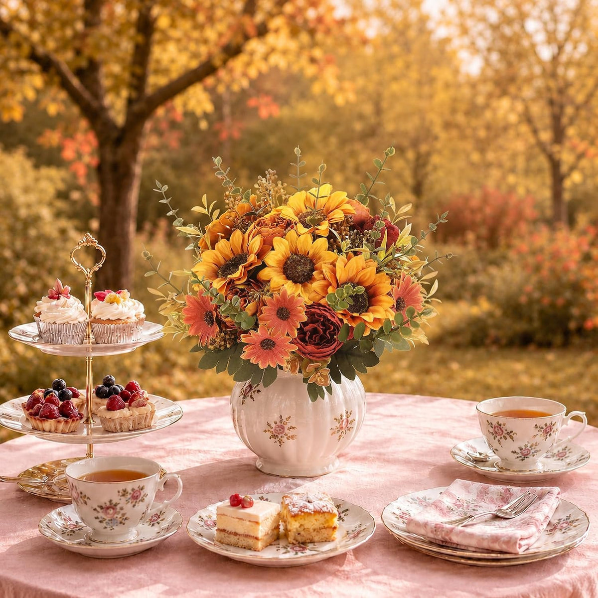 An autumn-themed floral centerpiece in a white floral vase sits on a pink tablecloth, surrounded by a tea and cake spread. The background shows a softly blurred outdoor park scene with golden fall trees.