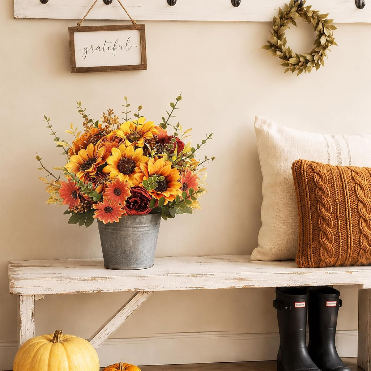 An autumn floral arrangement in a galvanized metal bucket sits on a rustic white bench. The bouquet features yellow and orange sunflowers, burgundy roses, and green foliage.