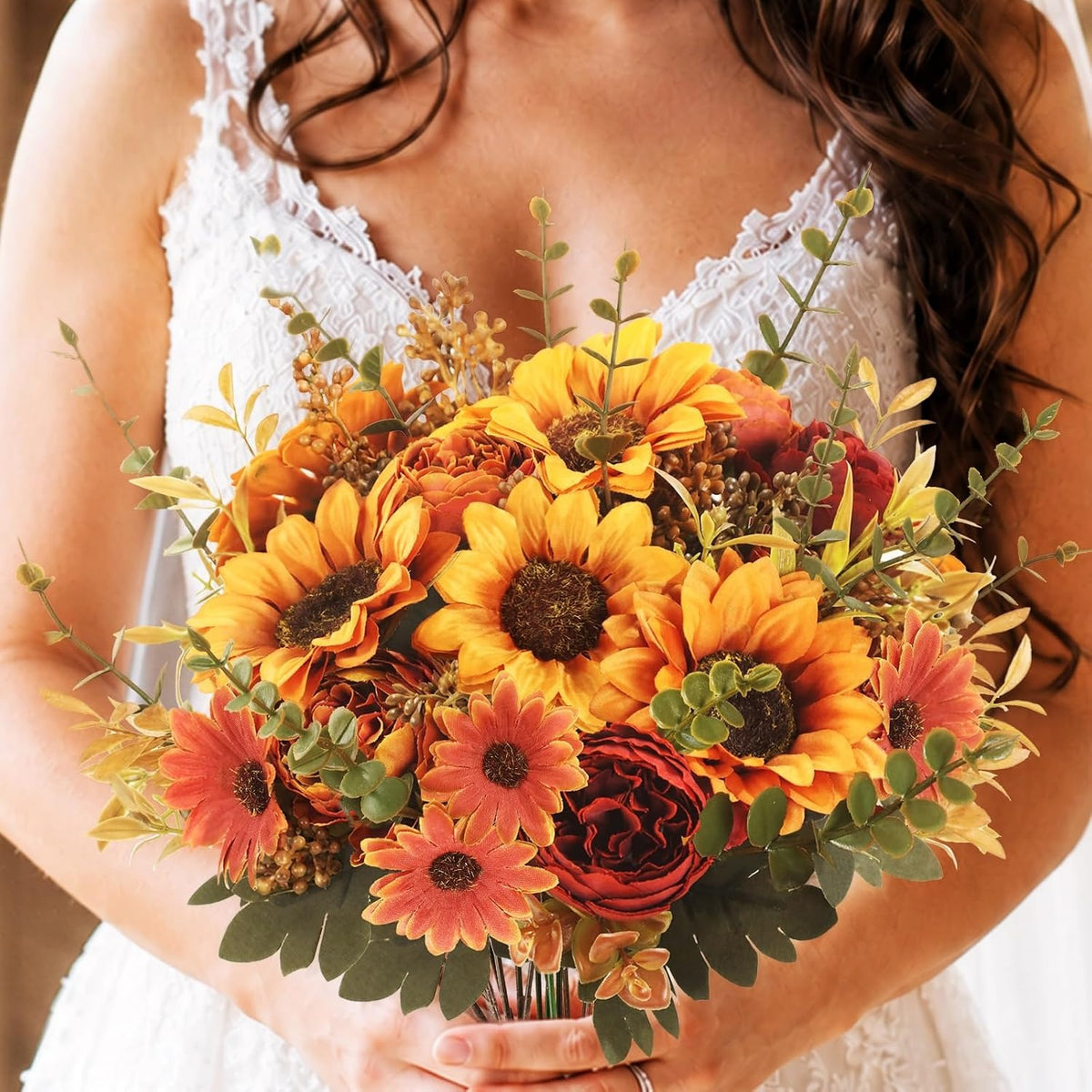 A close-up of a person in a white lace dress holding a vibrant artificial flower bouquet featuring sunflowers, orange daisies, burgundy roses, and green eucalyptus.